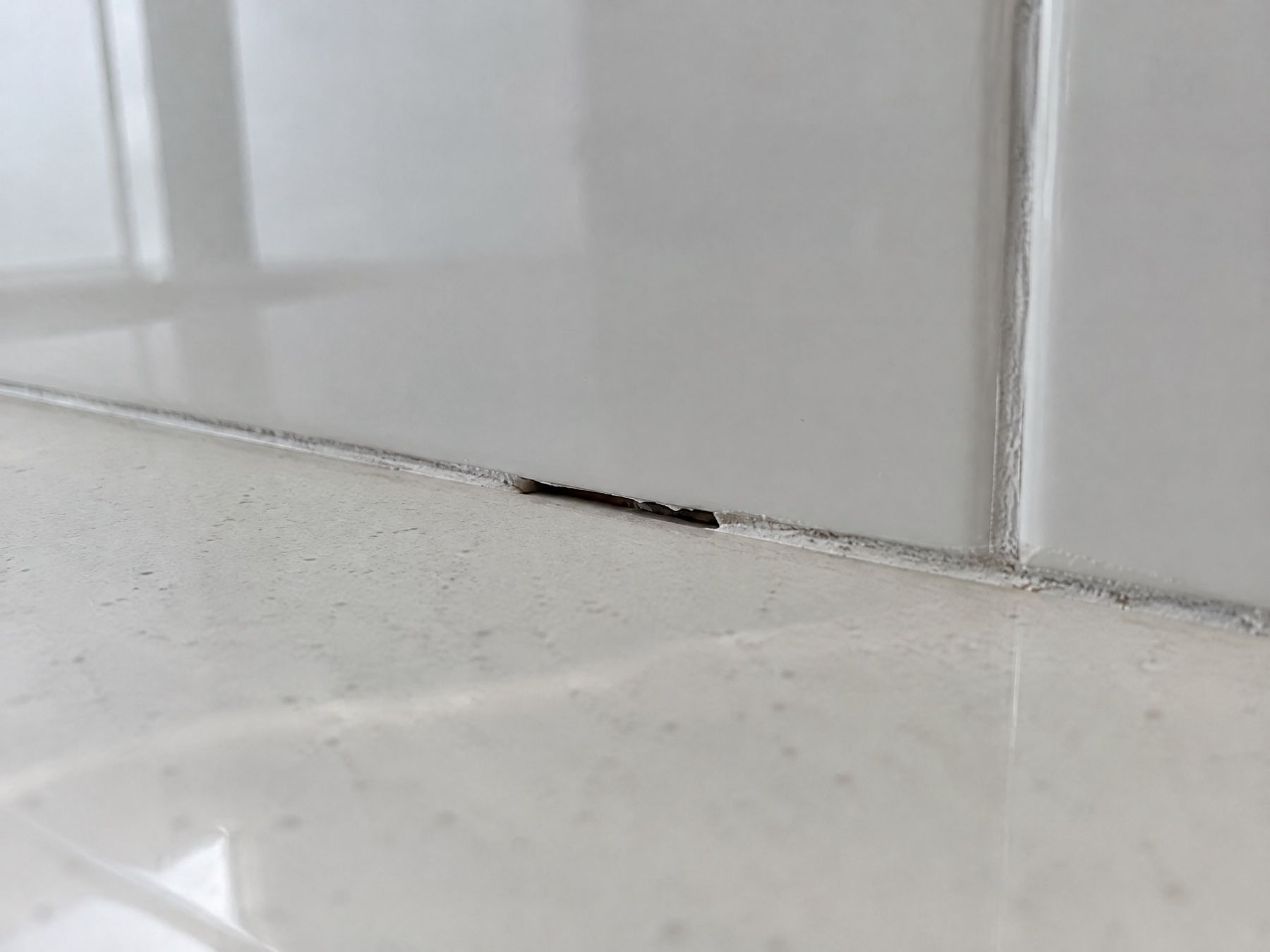 Close-up of a 3 mm gap in the grout line where the white wall tiles meet the floor tiles, circled in red.